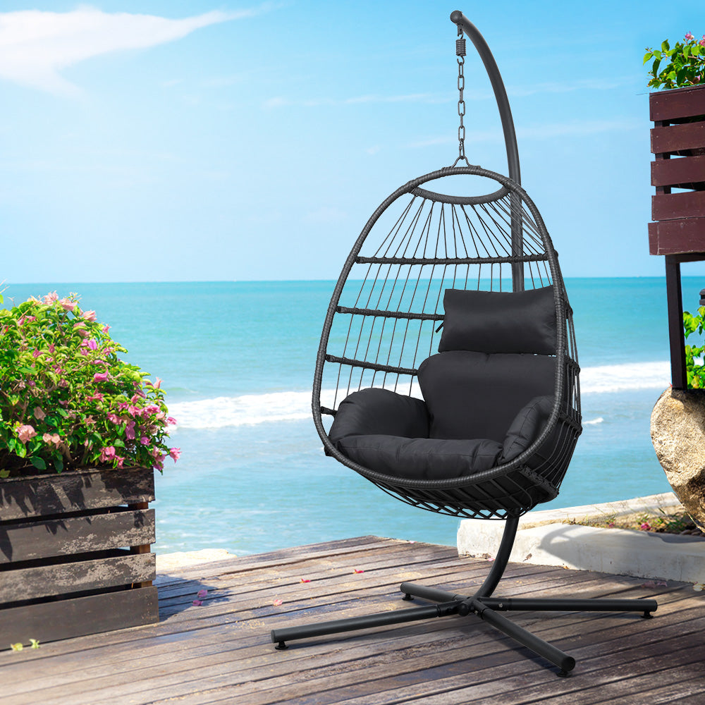 Black hanging egg chair with cushions on wooden deck overlooking ocean, perfect for outdoor relaxation.